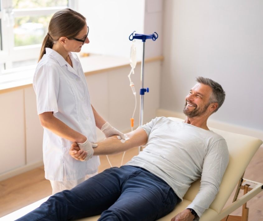 Smiling nurse attending to a man receiving an IV drip while lying on a hospital bed.