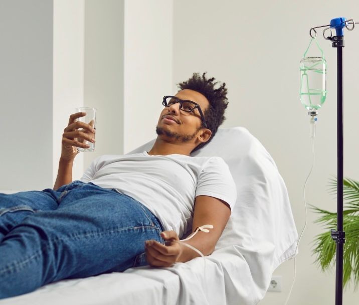 Person lying on a hospital bed with an IV drip and holding a glass of water.