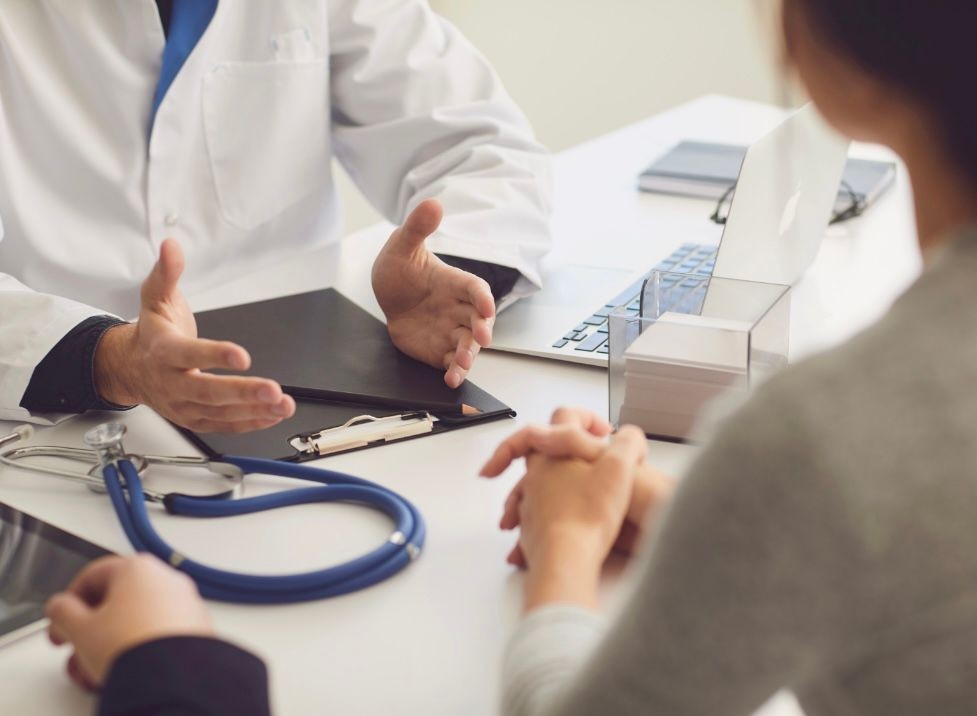 Doctor in white coat discussing with patient at a desk, stethoscope and clipboard visible.